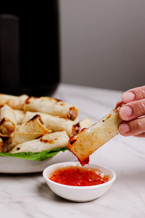 Air fryer frozen spring rolls recipe bite shot, dipping a spring roll in a traditional sweet and spicy sauce.