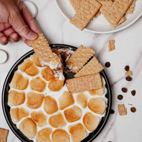 Air fryer s'mores dip recipe bite shot served on a pizza pan, with graham crackers on the side and a hand holding a dipped graham cracker.