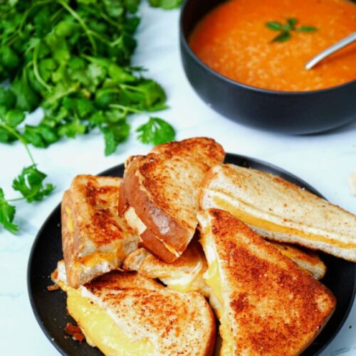 Air fryer grilled cheese sandwich recipe bite shot, stacked on a black plate with tomato soup.