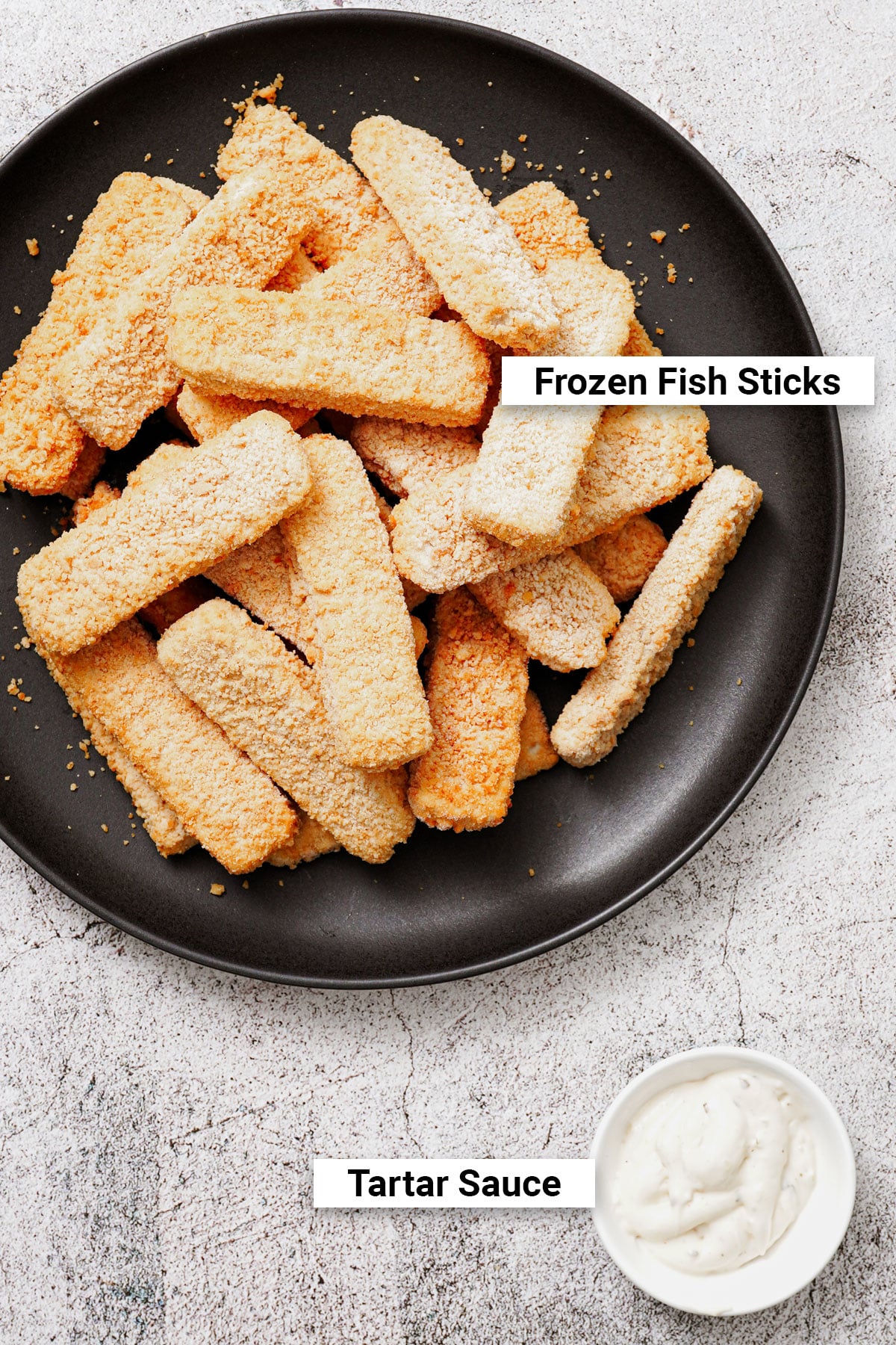 Uncooked frozen fish sticks on a black plate and tartar sauce in a dipping bowl.