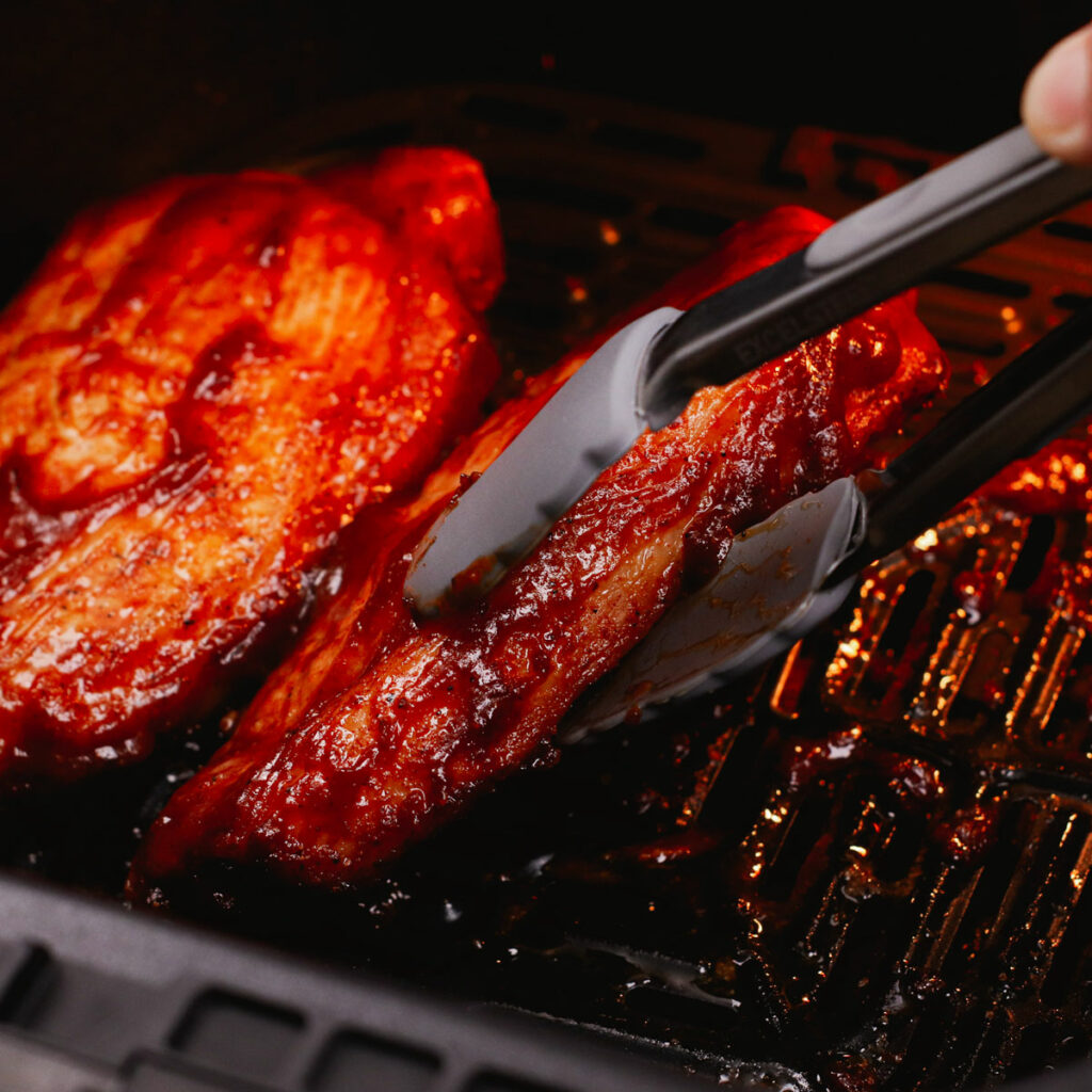 Flipping BBQ-flavored pork chops in the air fryer using a pair of tongs.